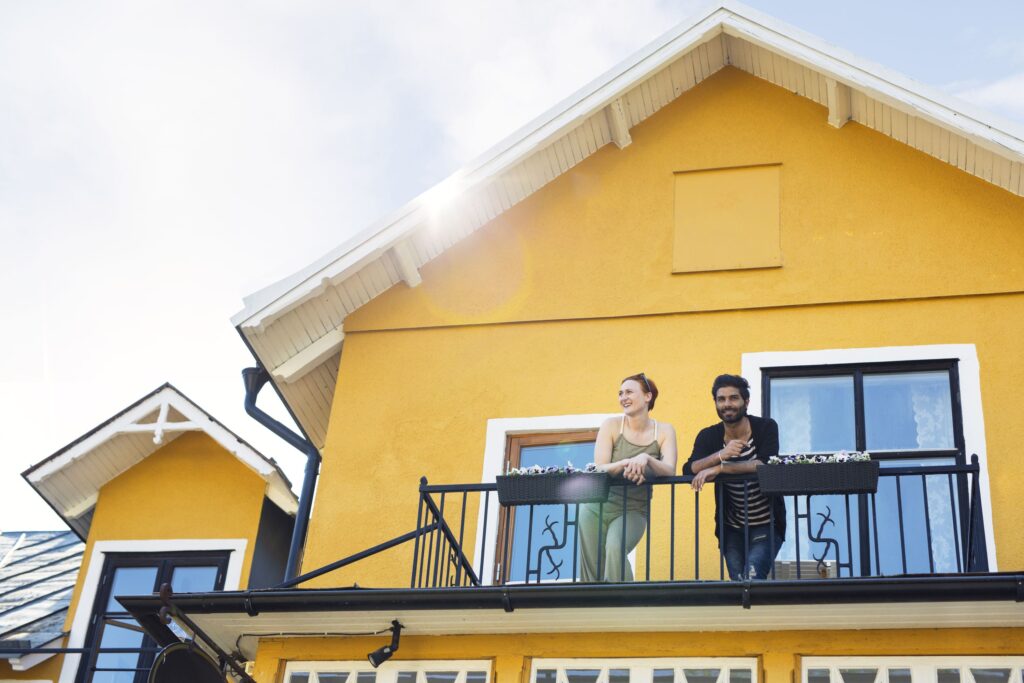 Eine Frau und ein Mann auf dem Balkon ihres gelben Hauses lächeln in die Kamera.