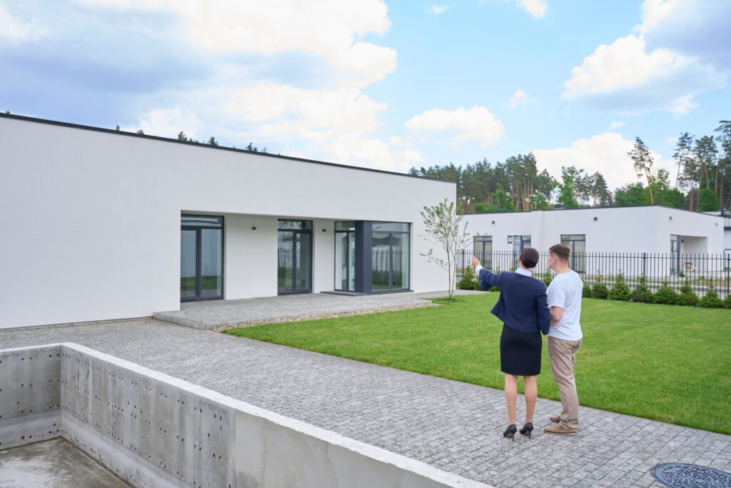 Abnahmefiktion im Bauvertragsrecht Hausbau Helden Eine Frau im Business-Outfit steht mit einem Mann in legerer Kleidung vor einem Haus. Sie zeigt darauf und beide besprechen etwas.
