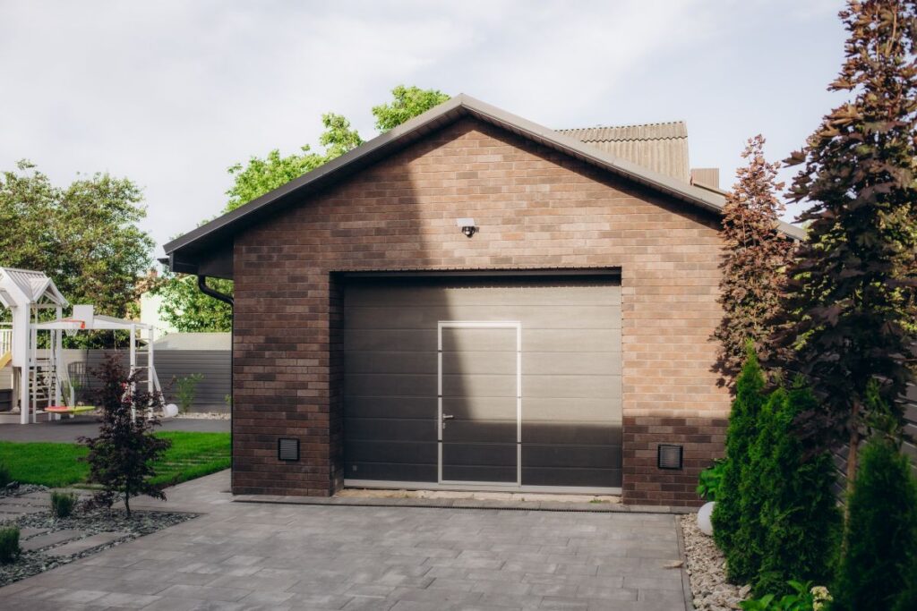 Garagenbau: Alles Wichtige von der Idee bis zur Fertigstellung Hausbau Helden Backstein-Garage mit grauem Garagentor umgeben von Garten mit Spielplatz für Kinder