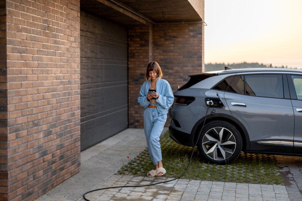 Garagenbau: Alles Wichtige von der Idee bis zur Fertigstellung Hausbau Helden Junge Frau benutzt Smartphone, während sie ihr Elektroauto in der Nähe der Garage ihres Hauses auflädt. Konzept des modernen Lebensstils und der Nachhaltigkeit