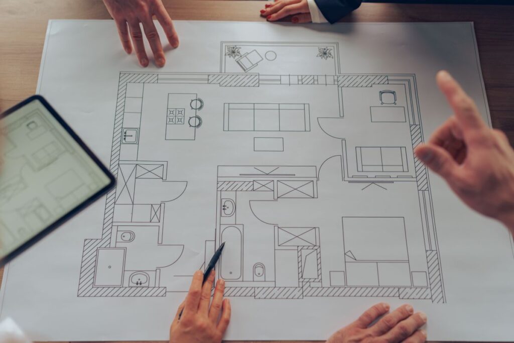 Günstiger Hausbau: Die 10 besten Tipps Hausbau Helden Grundriss einer Wohnung, einige Finger und Hände von Betrachtern sind dabei zu sehen