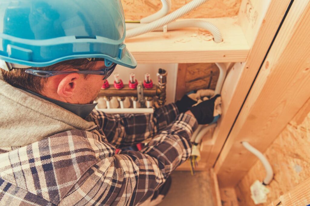 Ein Arbeiter mit blauem Schutzhelm und kariertem Hemd installiert vorsichtig Sanitärkomponenten in einem im Bau befindlichen neuen Gebäude.