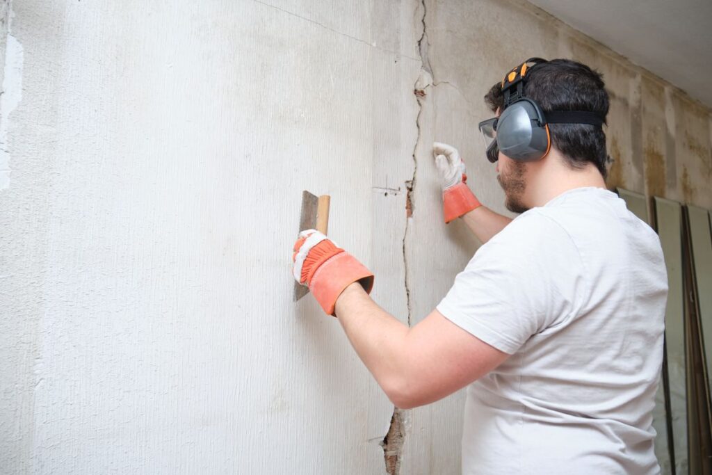 Bauarbeiter repariert einen Riss, verputzt Zement an der Wand. Baumeister trägt Schutzhandschuhe, Brille und Ohrenschützer.