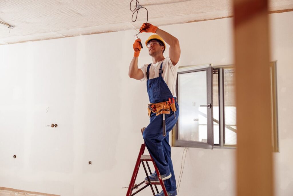 Die Elektrik eines Hauses und die Bedeutung von Qualität und Langlebigkeit Hausbau Helden Schöner junger Mann mit Schutzhelm steht auf einer Leiter und benutzt eine Zange, während er elektrische Leitungen repariert