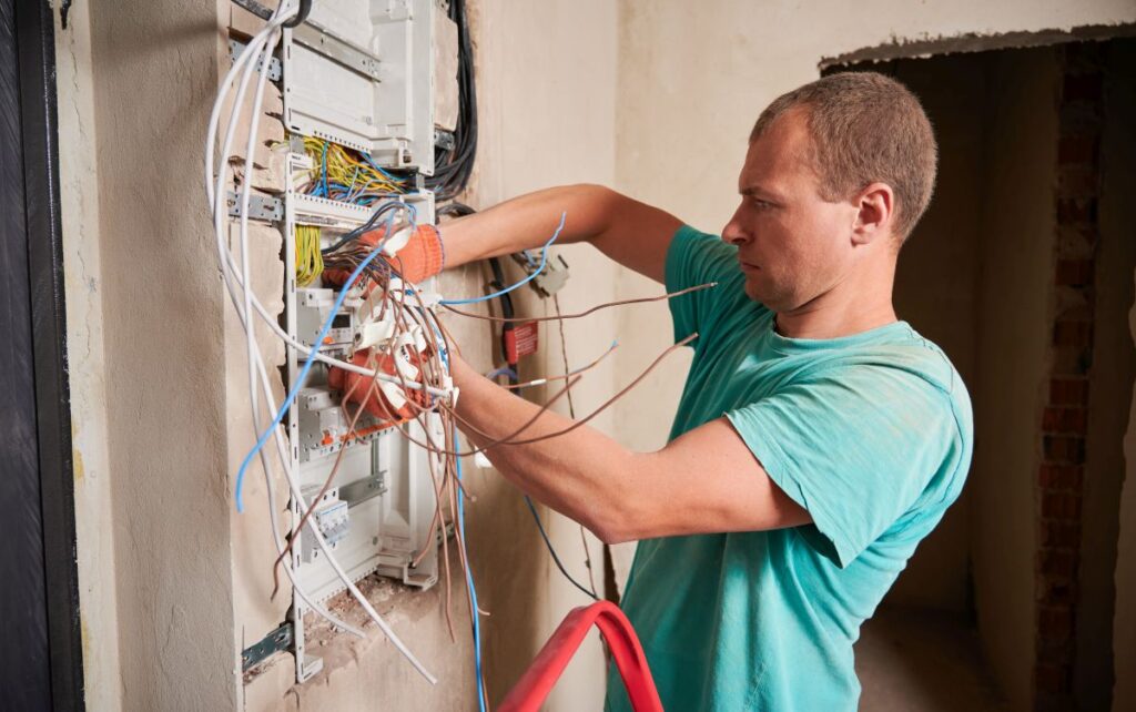 Seitenansicht eines Mannes in Arbeitshandschuhen, der die elektrische Schalttafel oder den Verteiler in einer renovierten Wohnung repariert. Elektriker repariert die elektrische Schalttafel mit Kabeln und Schaltern.